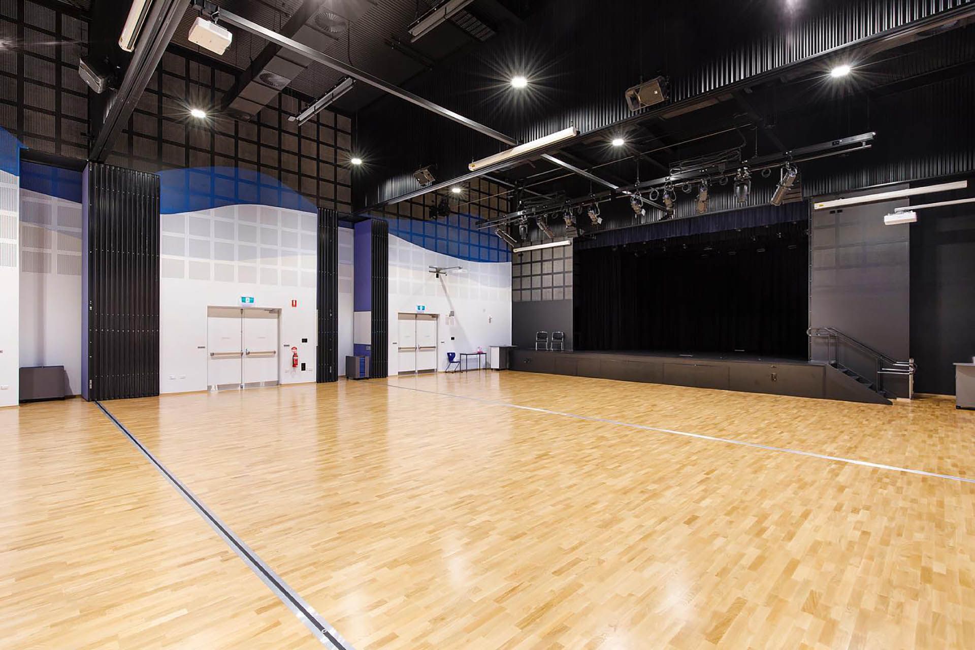 Empty indoor dance studio with polished wooden floor, black ceiling, and a stage along the back wall