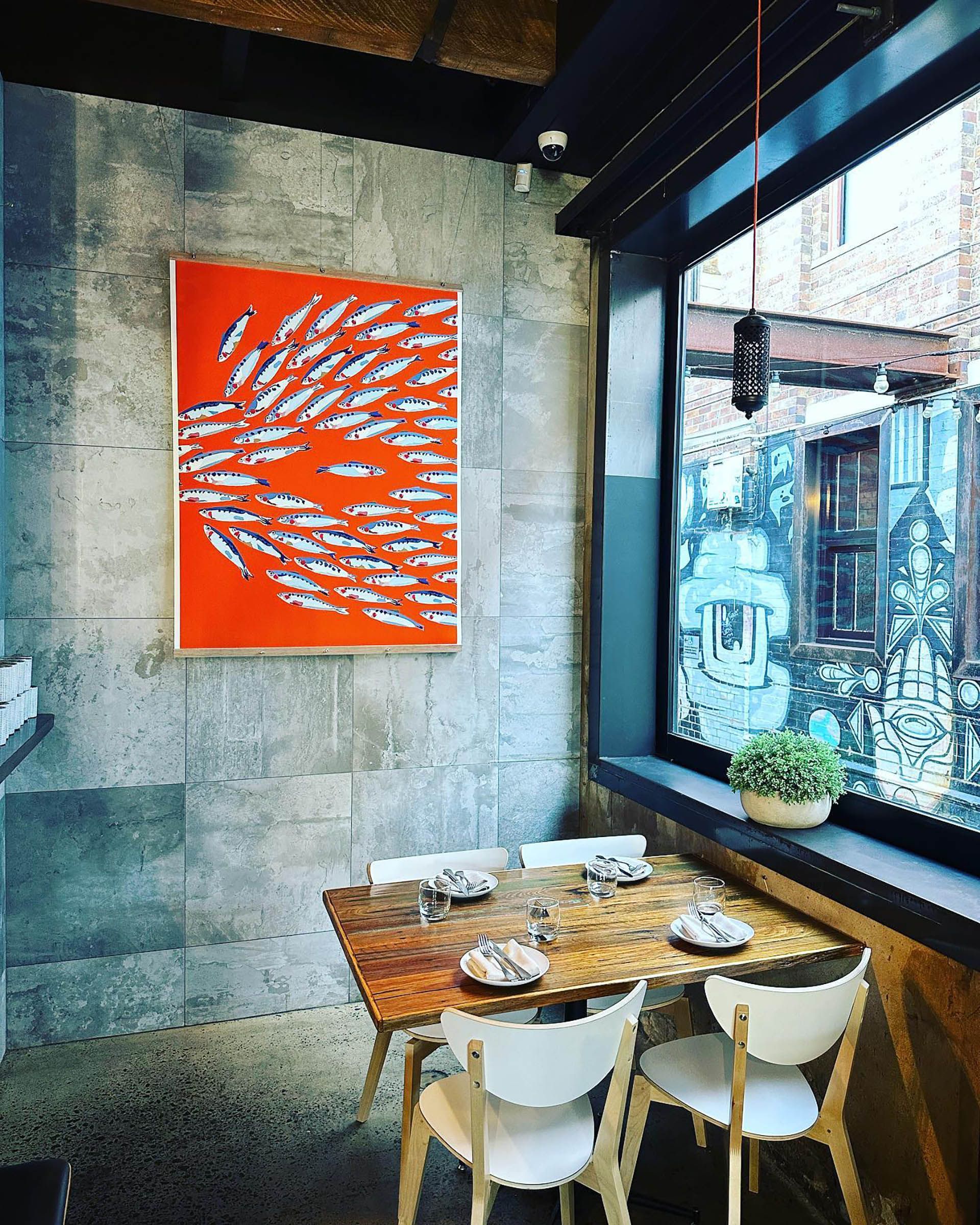 Cozy café corner with small wooden table, white chairs, orange wall art, and a large window