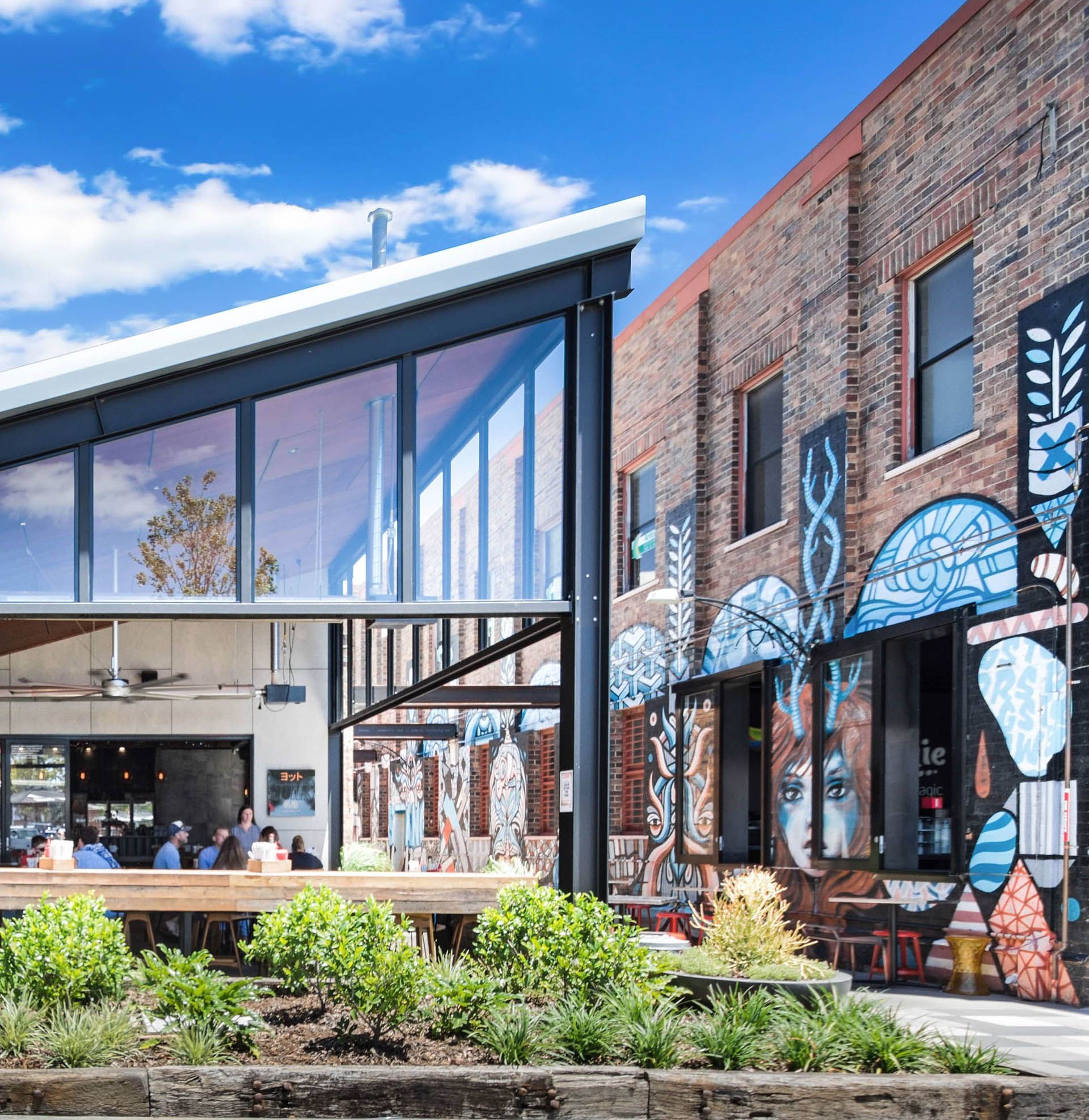 Modern brick storefront with colorful mural, glass patio enclosure, and outdoor seating under a blue sky