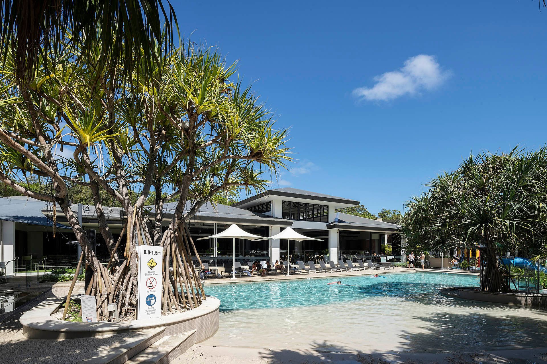 Outdoor resort pool with palm trees, lounge chairs, and a modern building under a blue sky