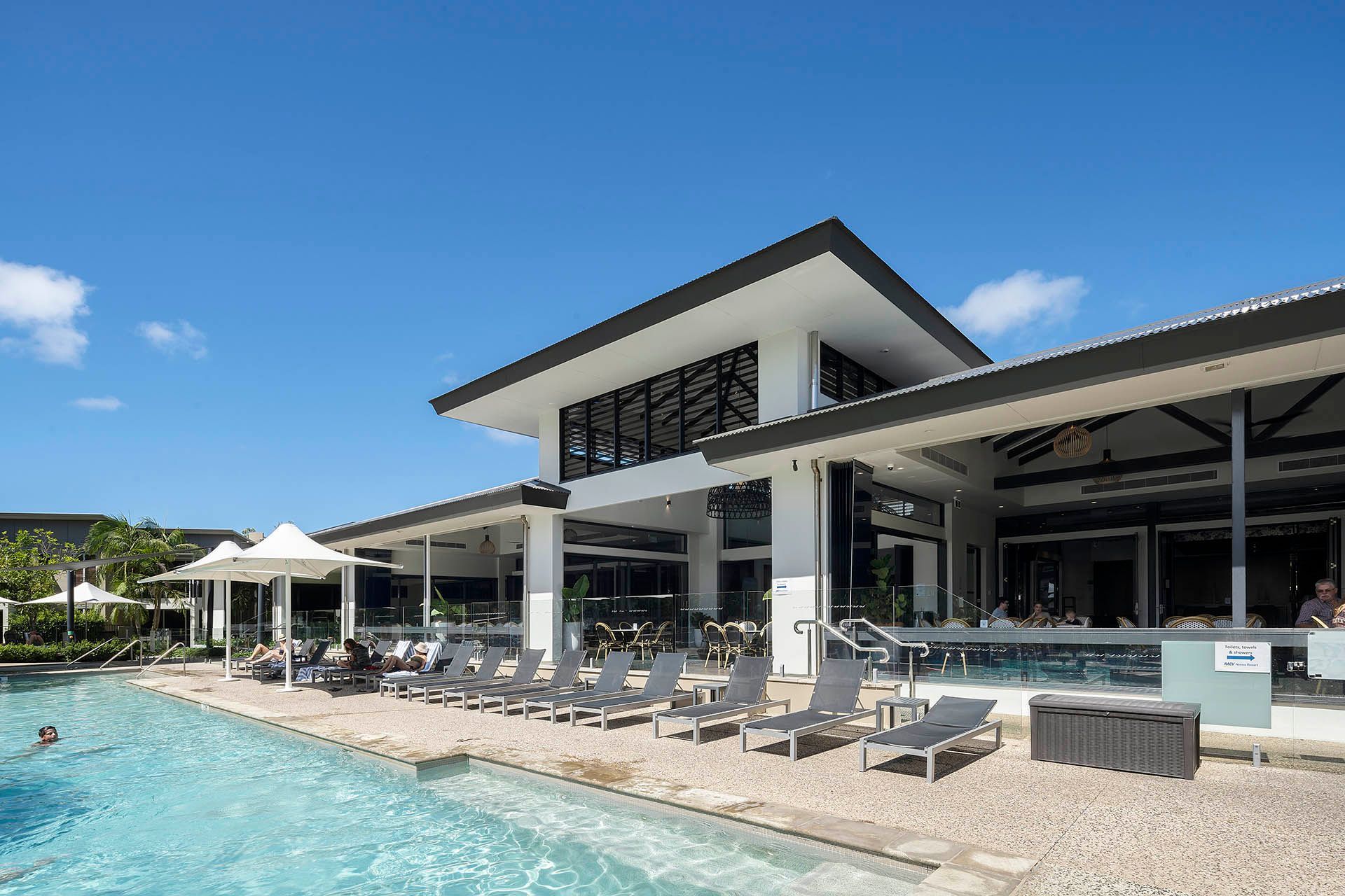 Modern resort poolside with lounge chairs, cabanas, and a large white pavilion by clear blue water