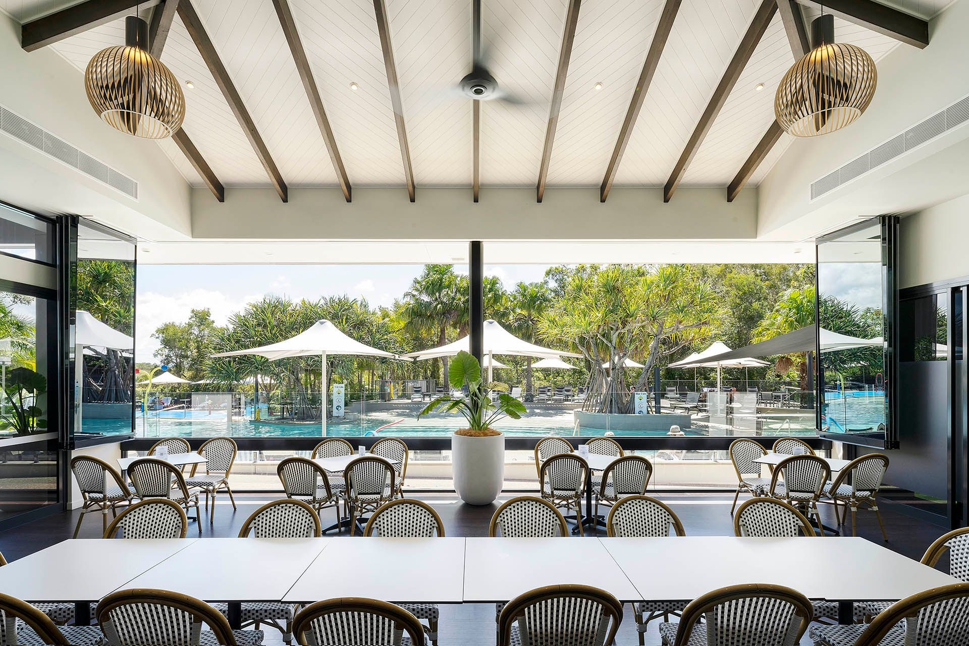 Open-air restaurant dining area with wicker chairs, tables, and a poolside view through large windows