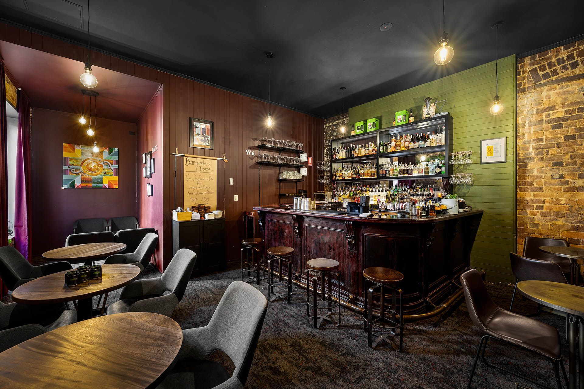 Cozy dimly lit bar interior with a wooden counter, stools, shelves of bottles, and small tables