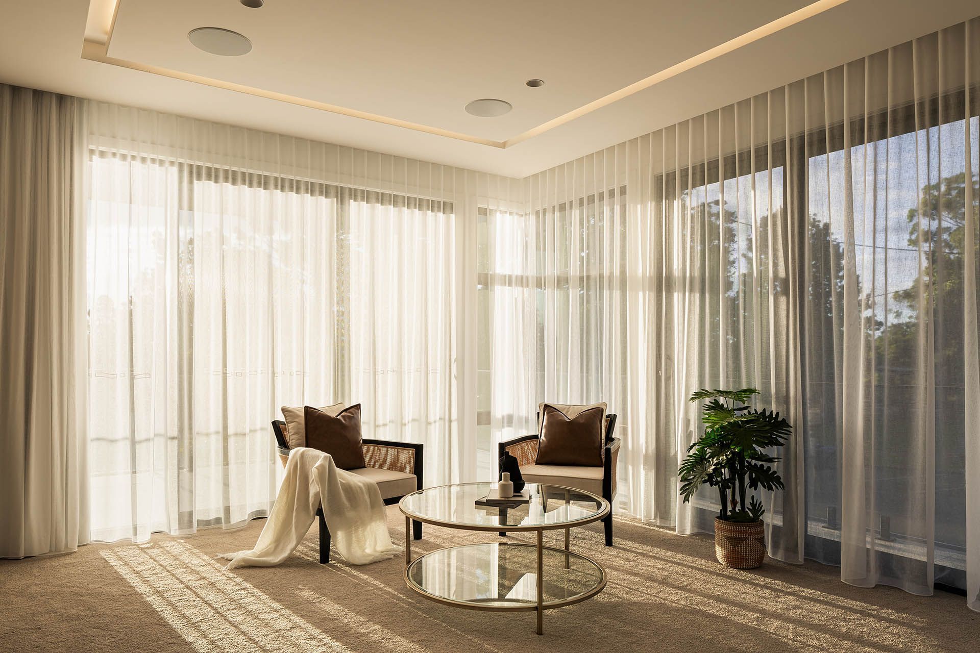 Sunlit living room with two armchairs, a side table, sheer curtains, and a potted plant by large windows.