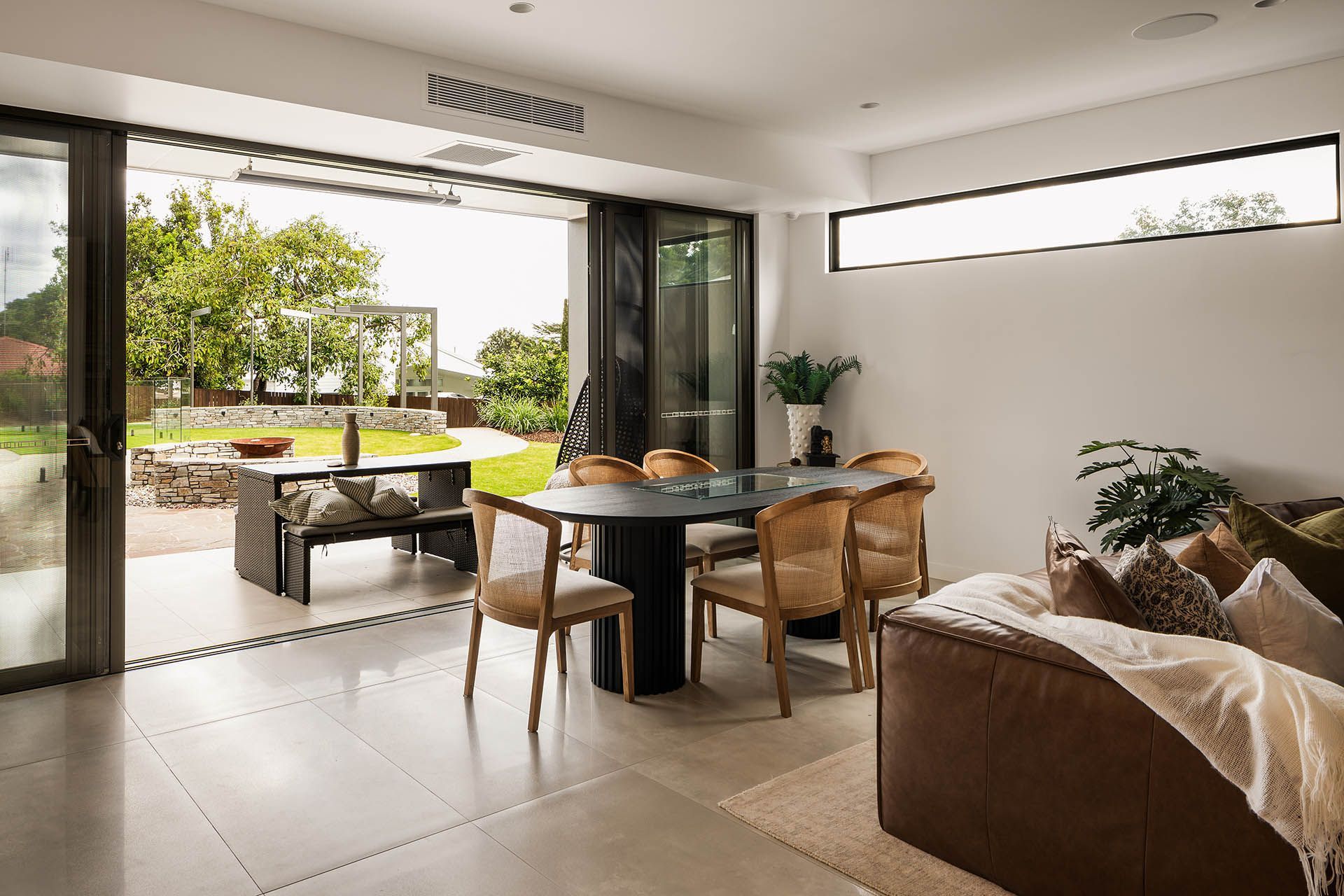 Modern living room opening to a bright patio and garden, with a round dining table and tan sofa
