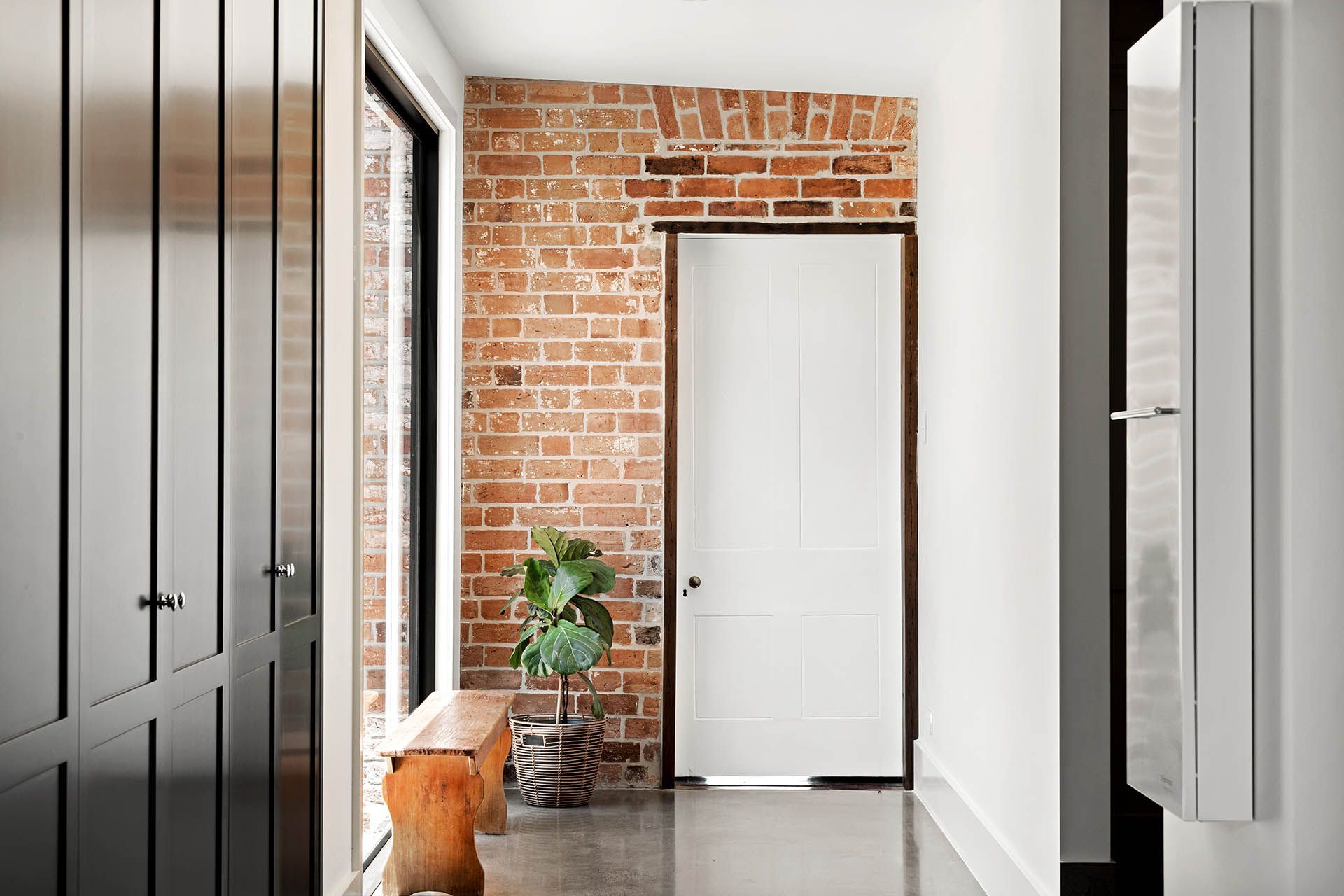 Narrow hallway with exposed brick wall, white door, and potted plant on a polished floor.