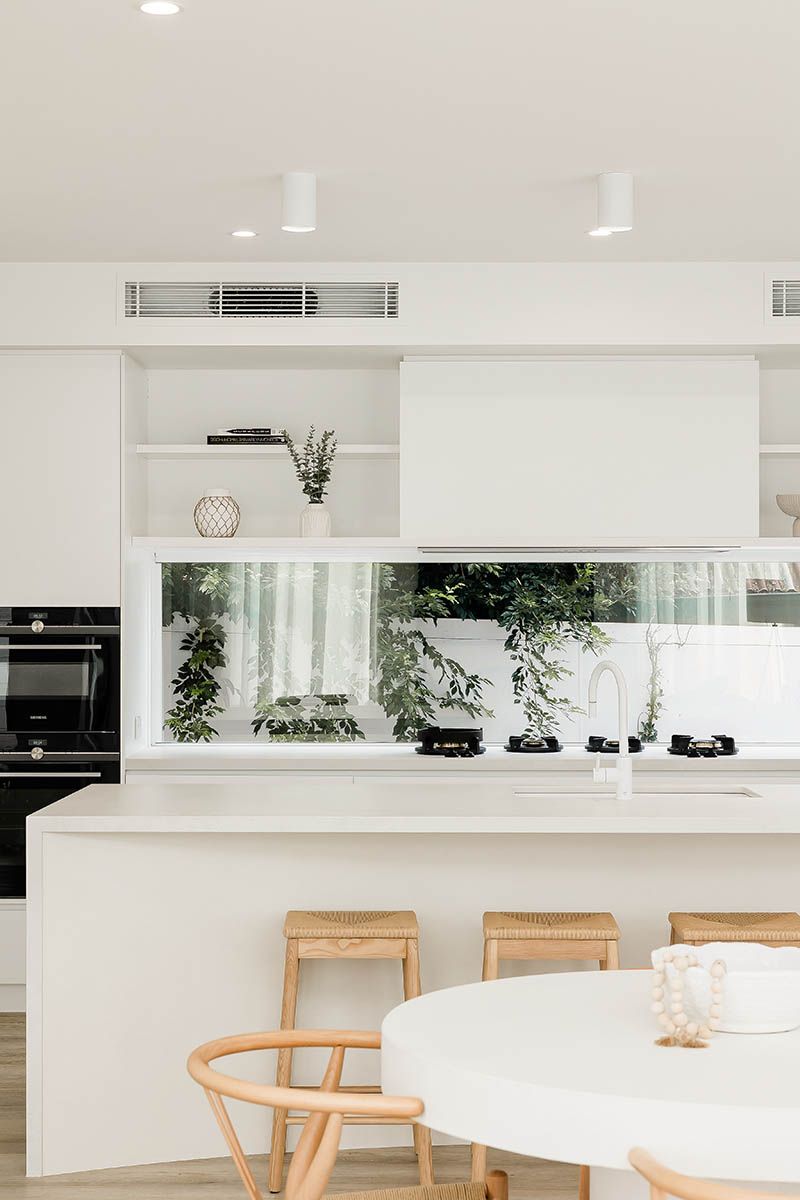 Bright modern white kitchen with an island, wooden stools, and a large window with greenery.