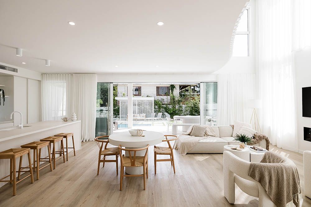 Bright, airy living-dining room with white decor, wooden table and stools, and large windows overlooking greenery