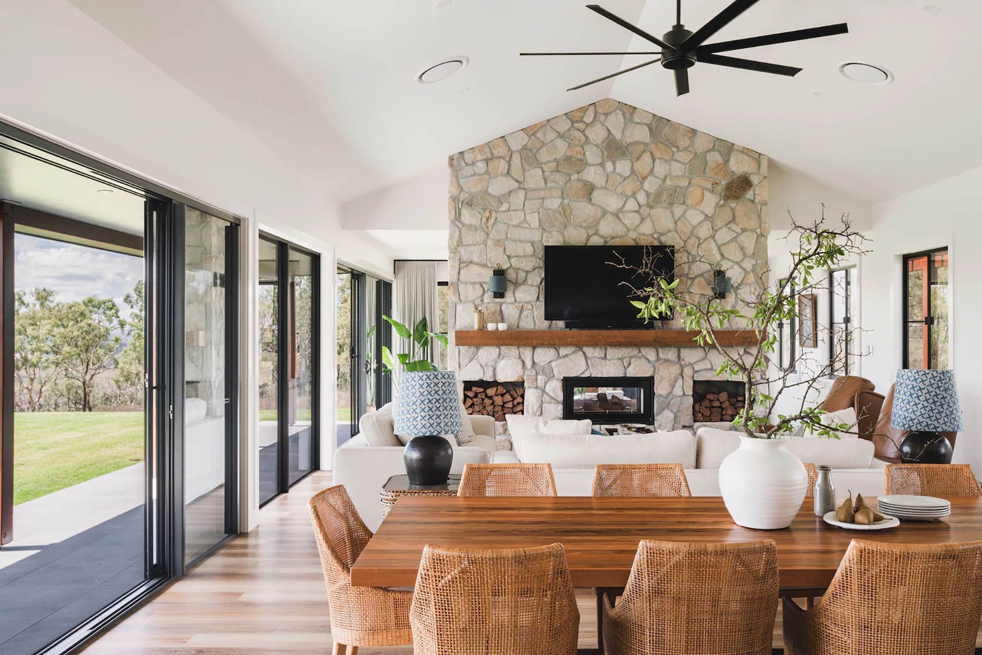Bright dining room with wicker chairs, stone fireplace, wall TV, and open glass doors to a yard
