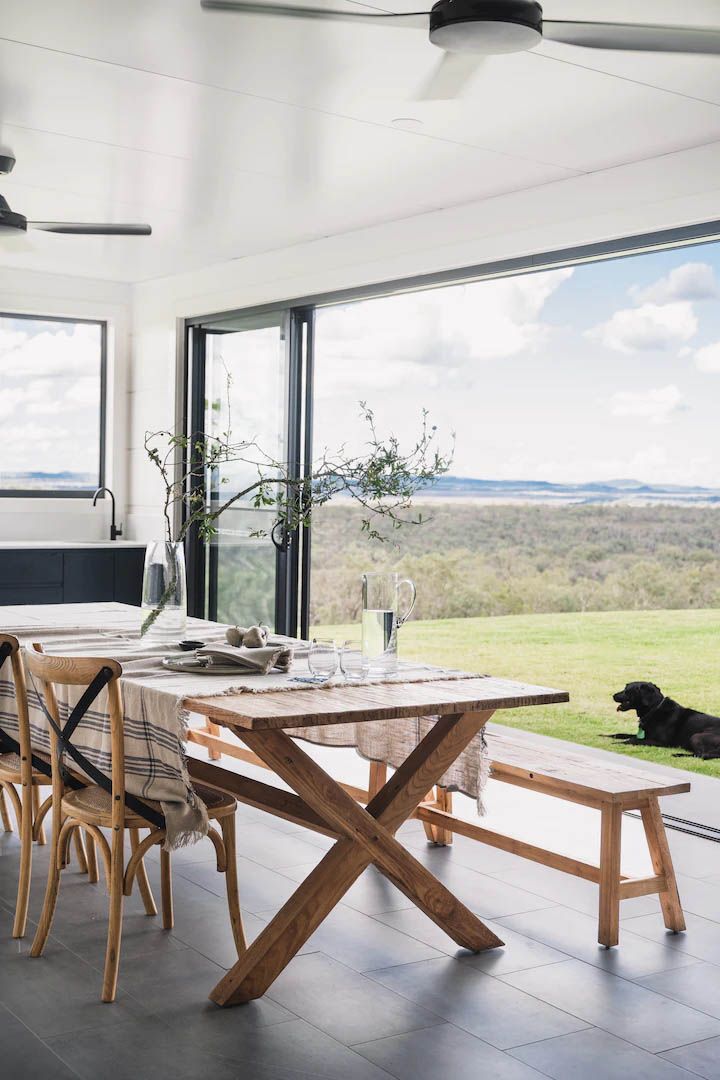 Bright dining room with wooden table and bench, open glass wall, and a black dog resting by the view.