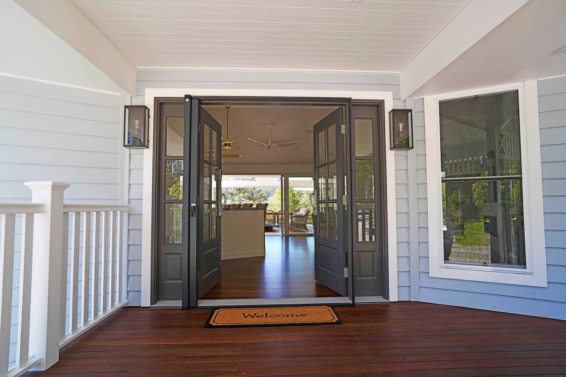 Covered porch with open double glass doors, wooden floor, white walls, and a doormat.