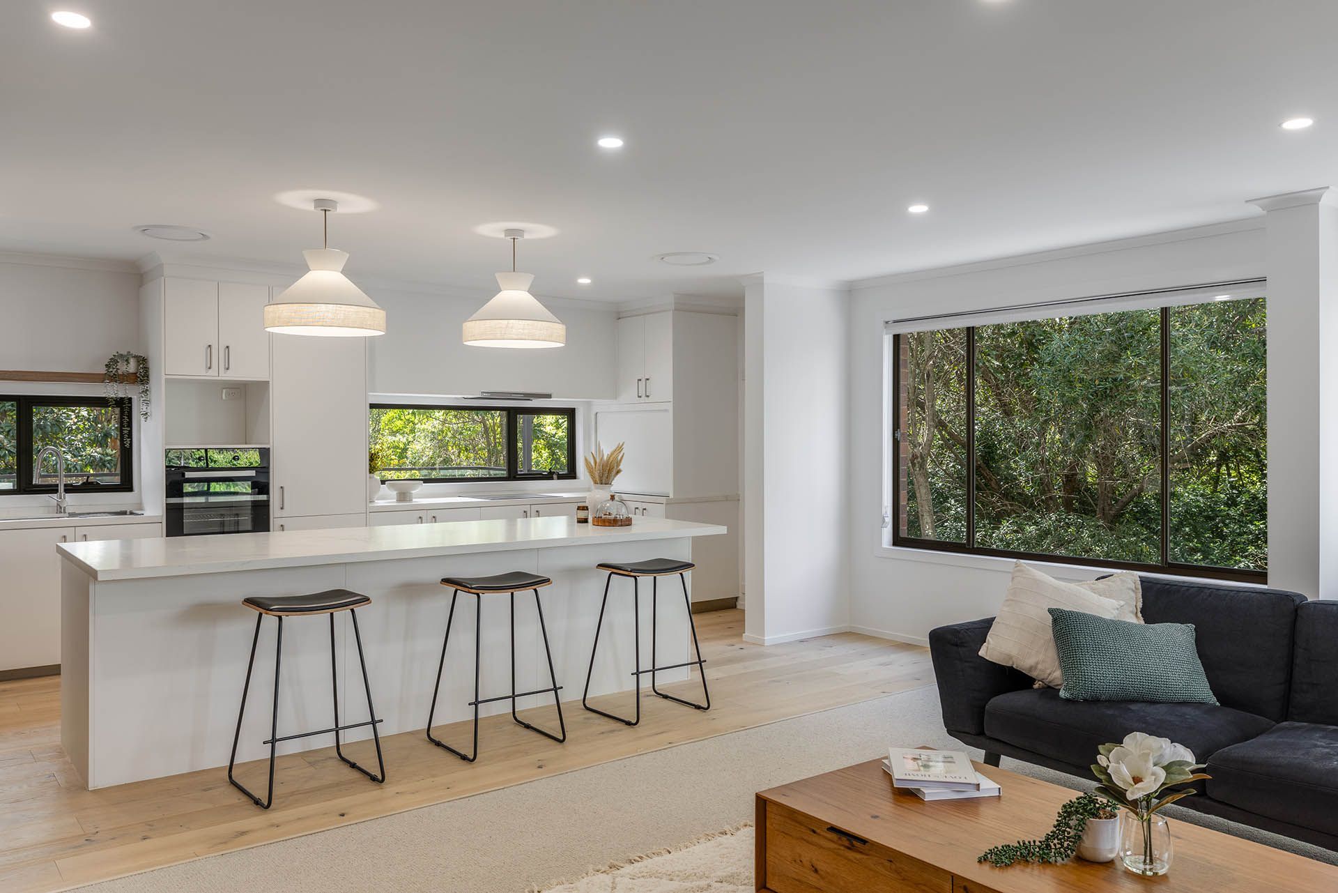 Bright modern kitchen and living room with white island, barstools, large windows, and dark sofa