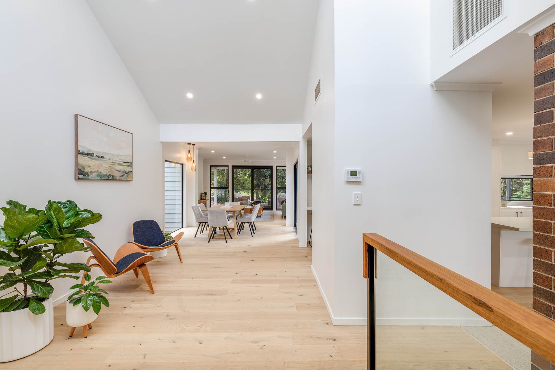 Bright modern hallway with light wood floors, white walls, plants, and a view to a dining area and glass doors.