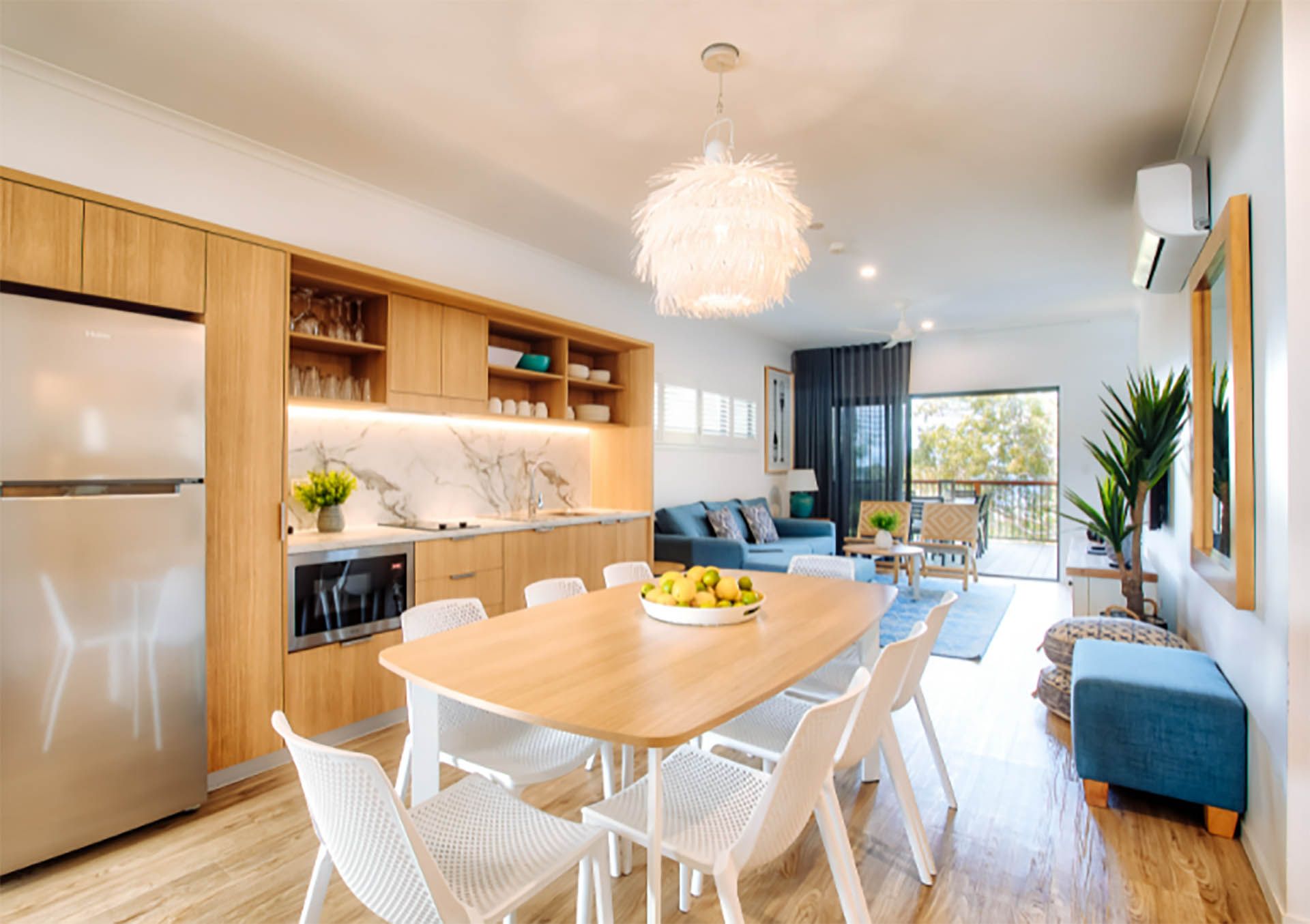 Bright modern kitchen-dining area with wood cabinetry, white chairs, and sunlight from large windows.