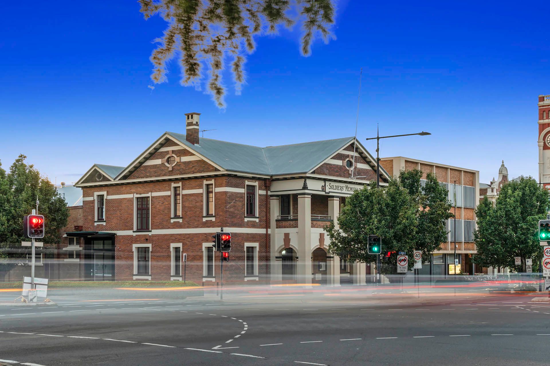 Historic brick building at a city intersection, with trees, traffic lights, and blurred cars on the street.