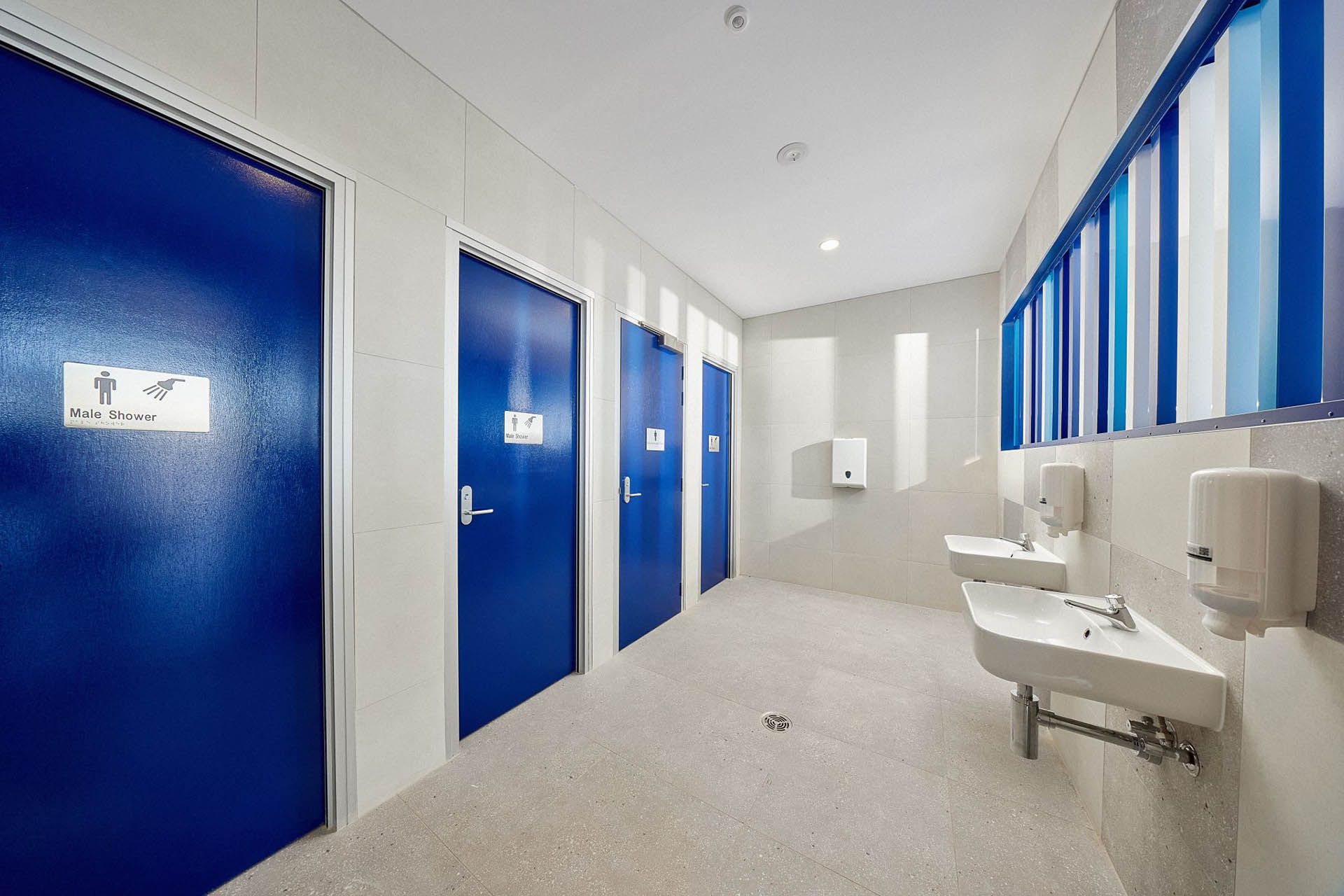 Bright restroom corridor with blue doors, white sinks, and striped windows on the right