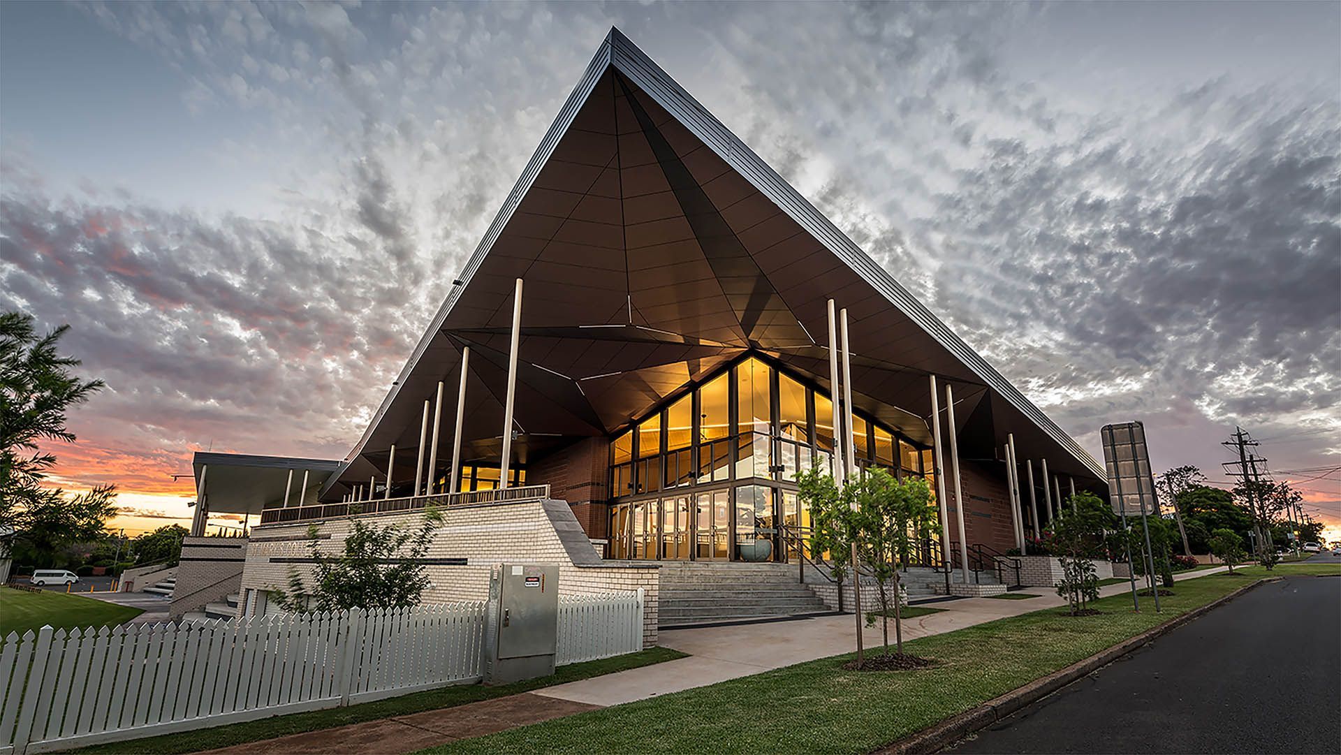 Modern A-frame building at dusk with warm interior lights, glass facade, and landscaped entrance