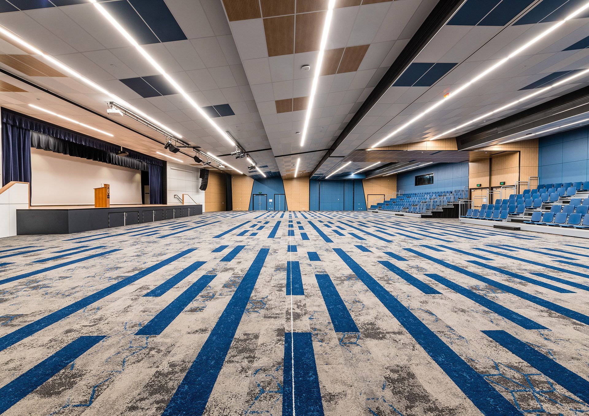 Empty conference hall with blue-and-gray carpet, rows of chairs, and a stage with curtains