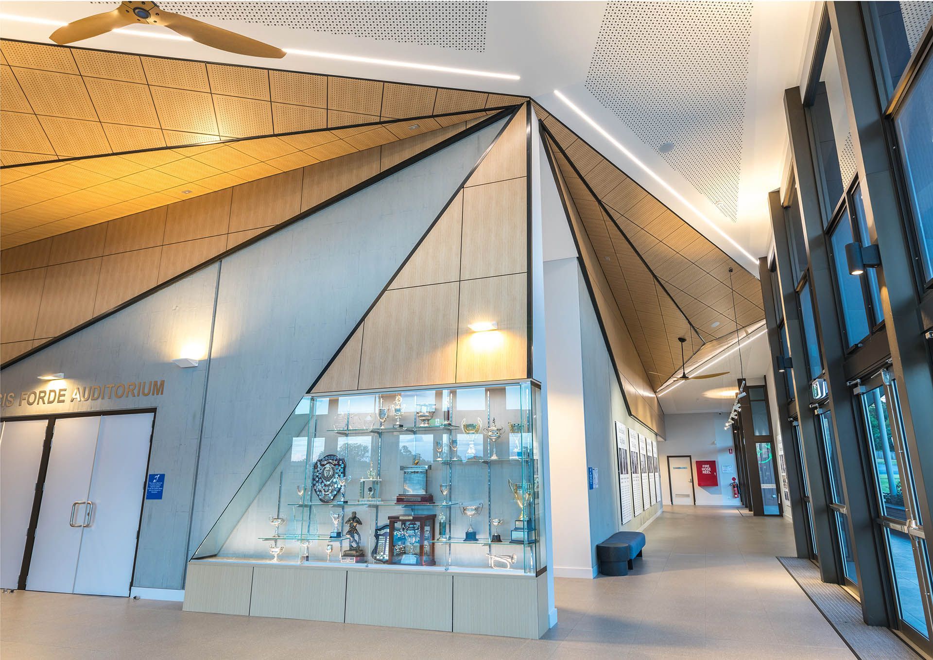 Modern indoor atrium with angular white-and-wood ceiling, glass display, and corridor seating area