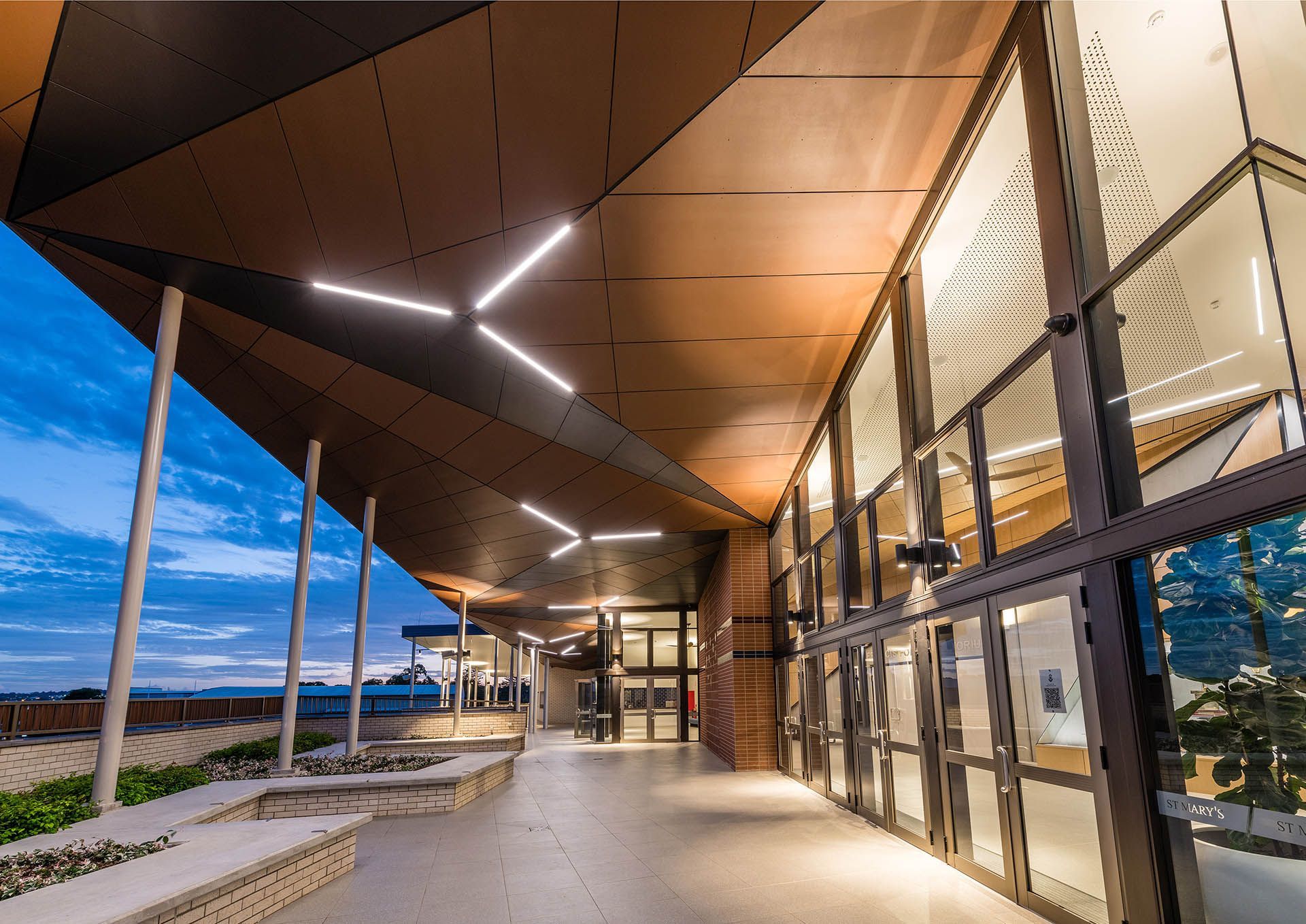 Modern building entrance with glass walls, wood ceiling, and geometric lights at dusk