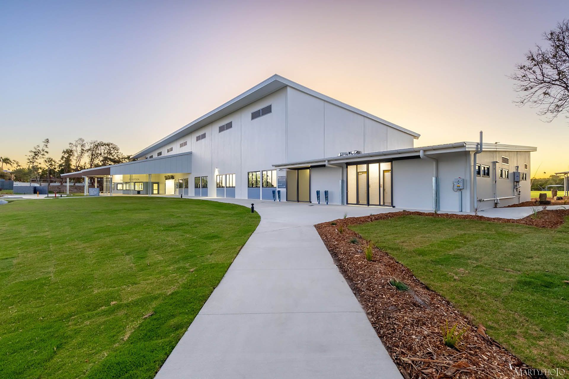 Modern white building with large windows, a walkway, green lawn, and sunset sky