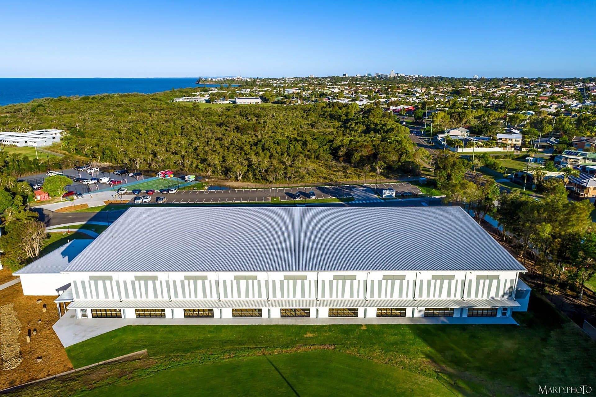 Aerial view of a large white-roofed building surrounded by green trees and a coastal neighborhood.