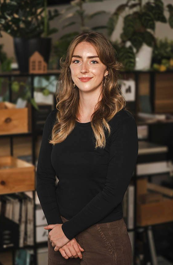 Portrait of a smiling woman standing indoors against shelves with plants and decor.