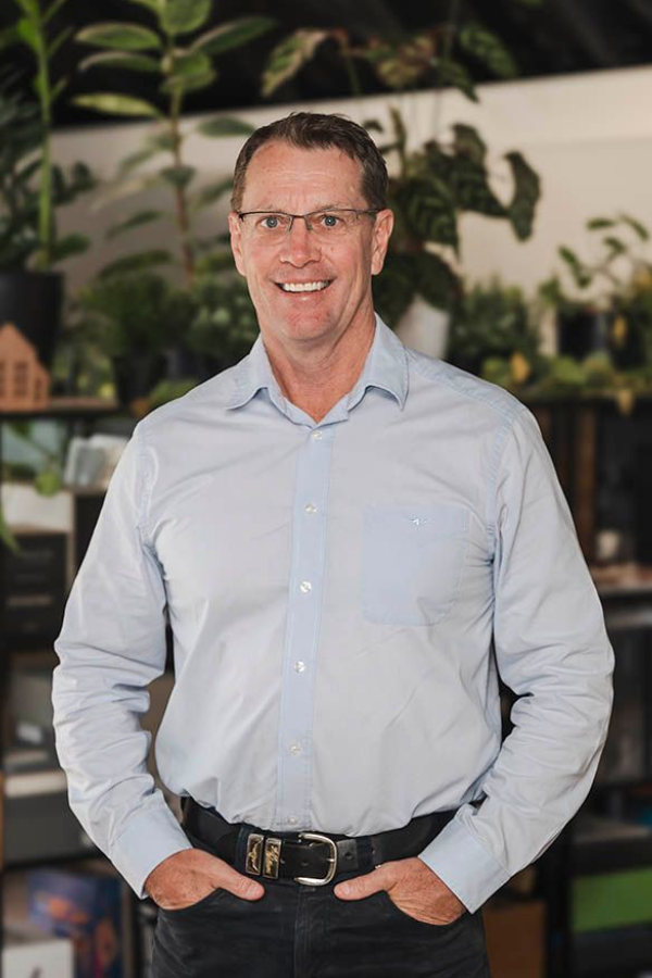 Smiling man in a light blue shirt standing indoors with hands in pockets, with plants in the background