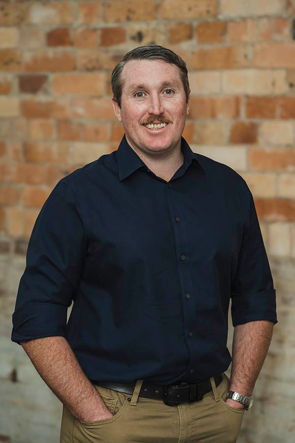 Man in a dark shirt standing against a brick wall, hands in pockets, smiling slightly