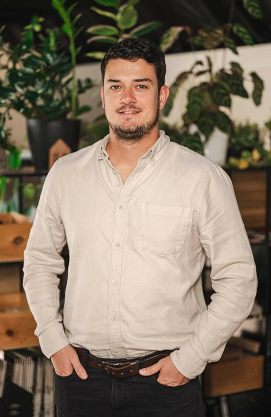 Man in a beige button-up shirt standing indoors with hands in pockets, in front of plants and shelves.
