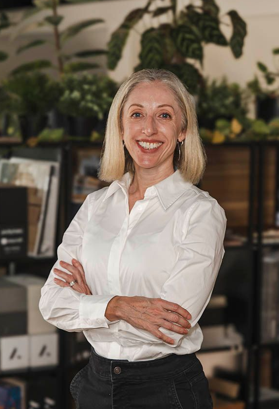 Smiling woman in a white shirt stands with arms crossed in a plant-filled office or shop.