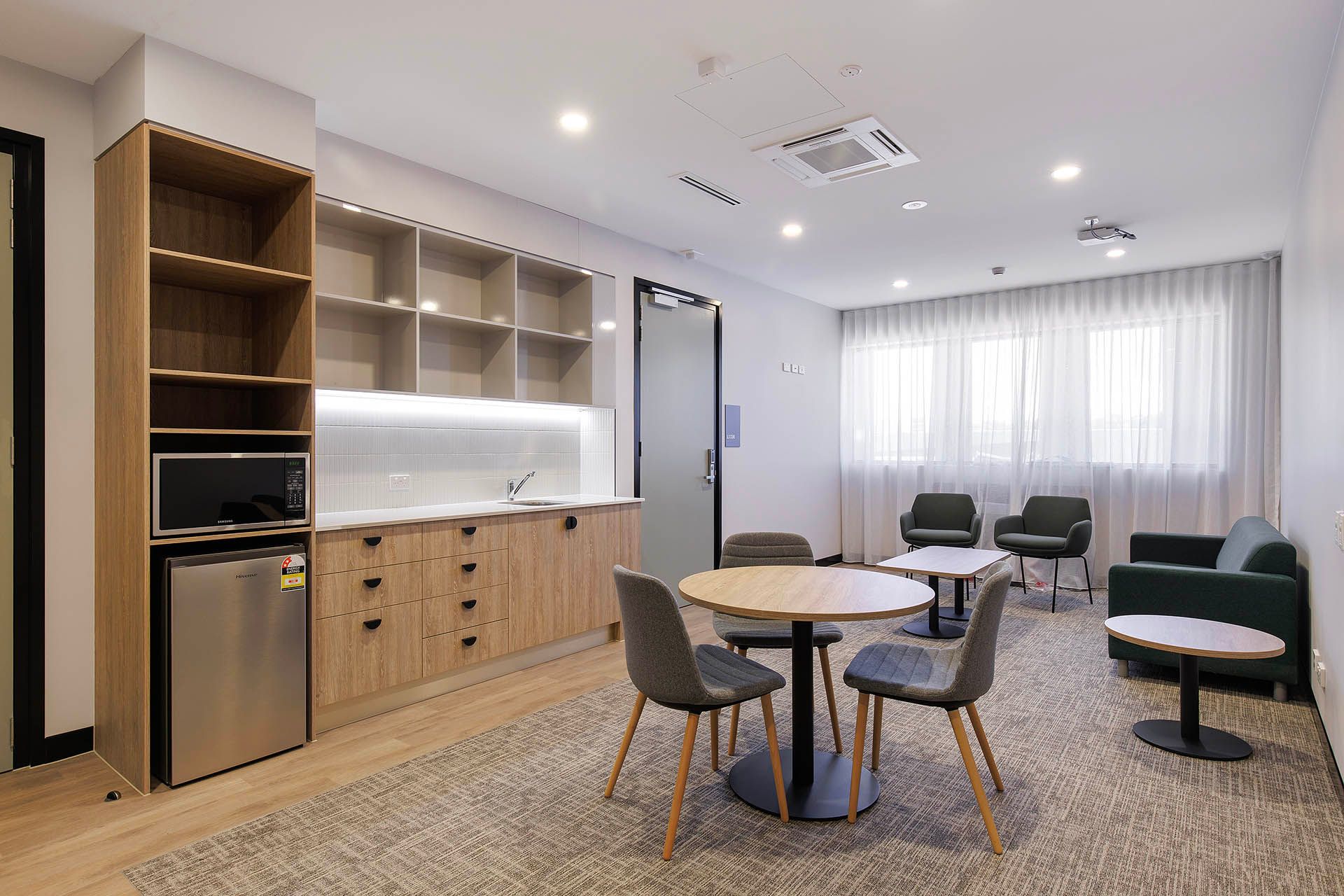 Modern lounge with kitchenette, round tables, chairs, sofas, and sheer curtains in a bright room.