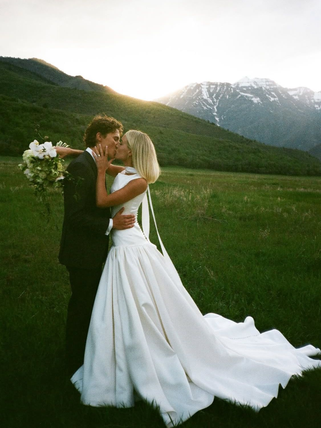 Couple kissing in wedding attire, embracing in a grassy field, with mountain backdrop at sunset.