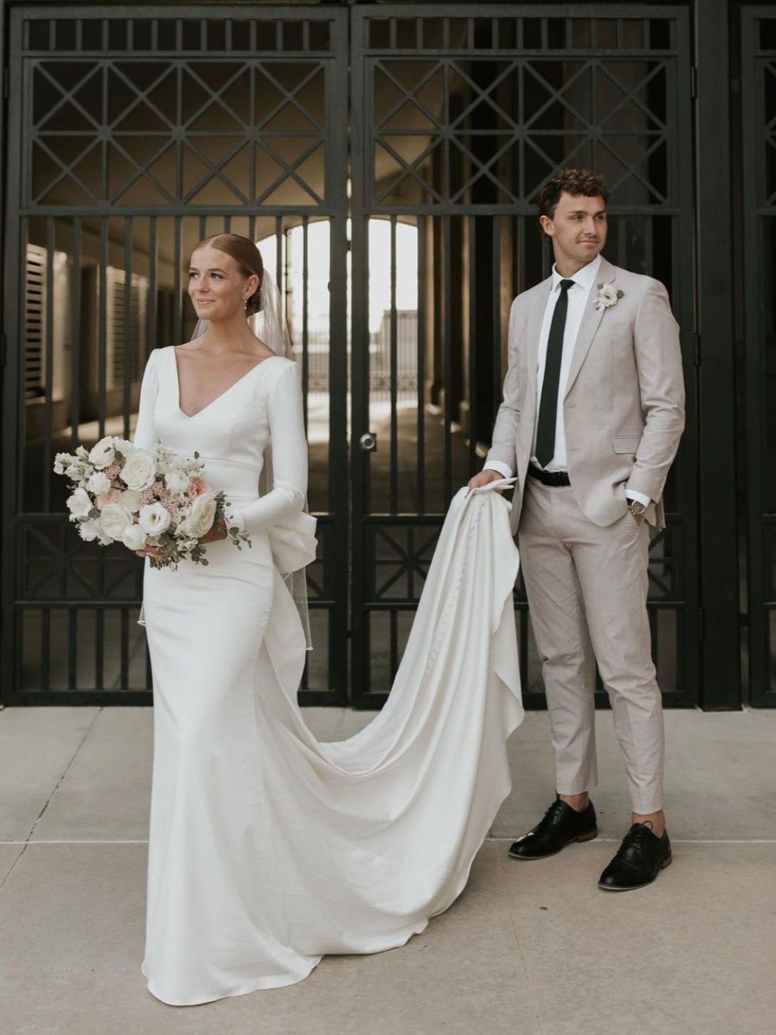 Bride in white dress with long train, groom in grey suit, standing by black metal gates.