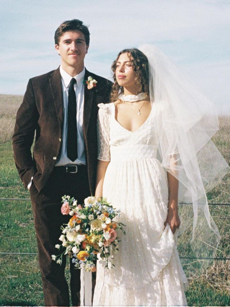Newlyweds pose in field: man in brown suit, woman in white dress and veil, holding bouquet.