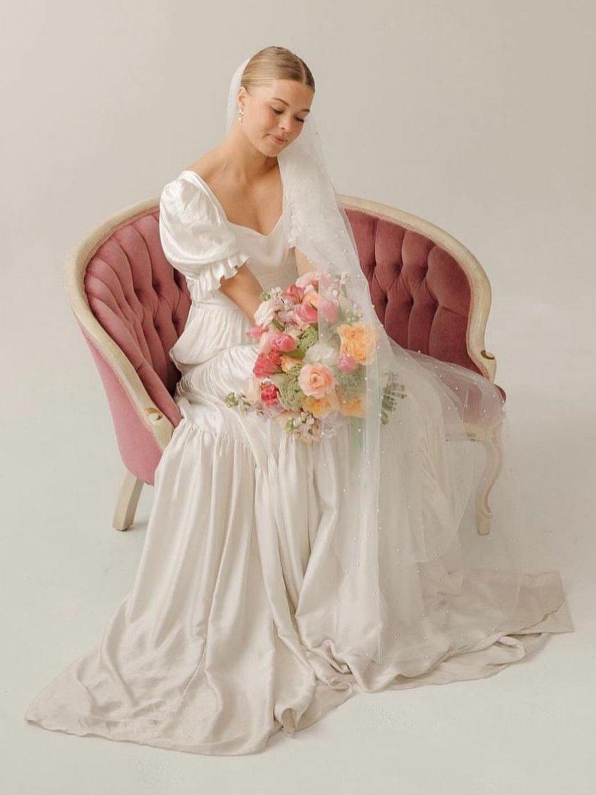 Bride wearing a wedding gown and veil, holding bouquet, seated on a pink antique chair.
