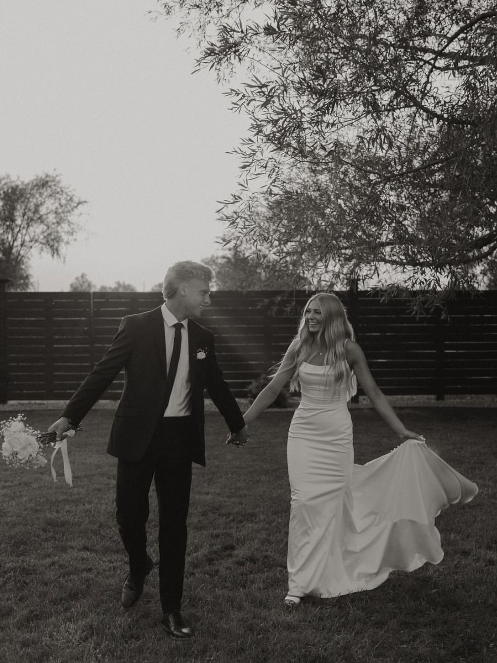 Bride and groom walk hand-in-hand in a grassy outdoor area, holding hands, her dress trailing, in black and white.