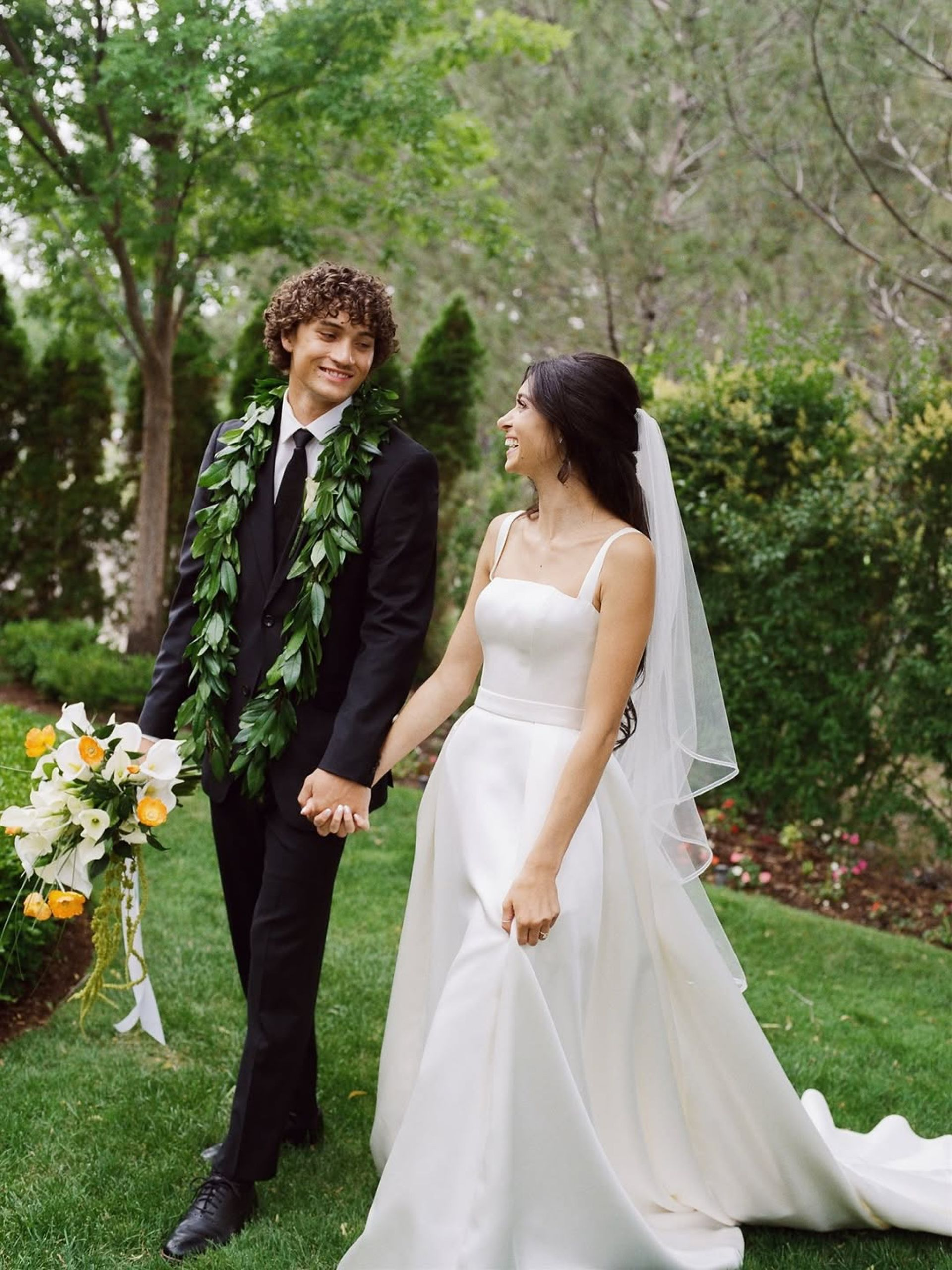 Couple holding hands, walking in a garden. The woman wears a white wedding dress and veil. The man wears a black suit