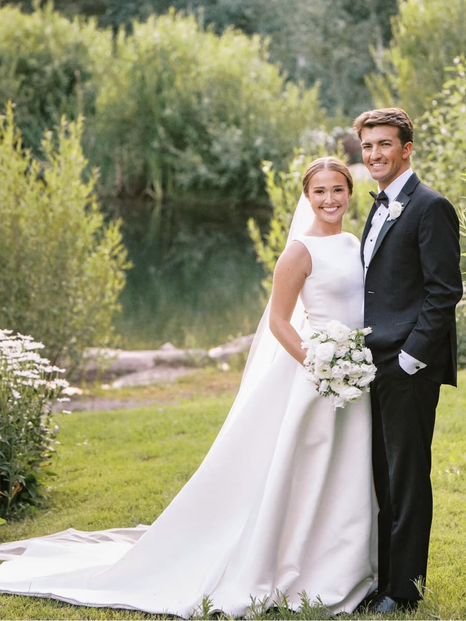Bride and groom pose outdoors; she in white gown, he in black tuxedo, smile at camera.