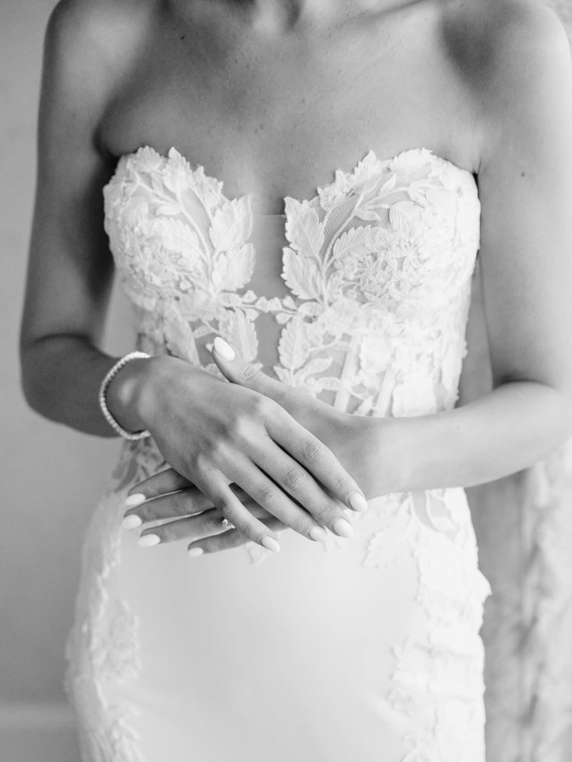 Woman wearing a strapless lace wedding dress, hands clasped, silver bracelet.