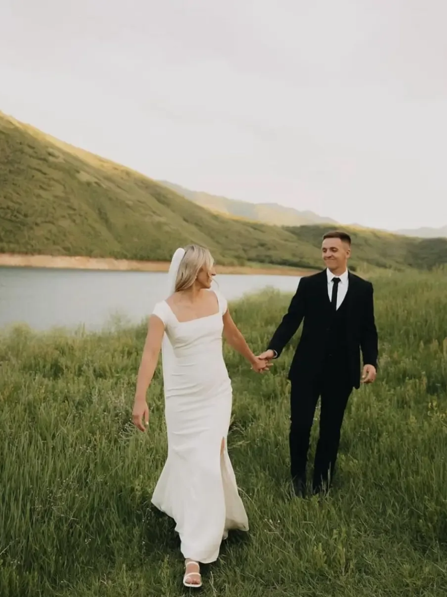 Bride and groom hold hands walking in a field. Bride in white dress, groom in black suit, lake and mountains in background.
