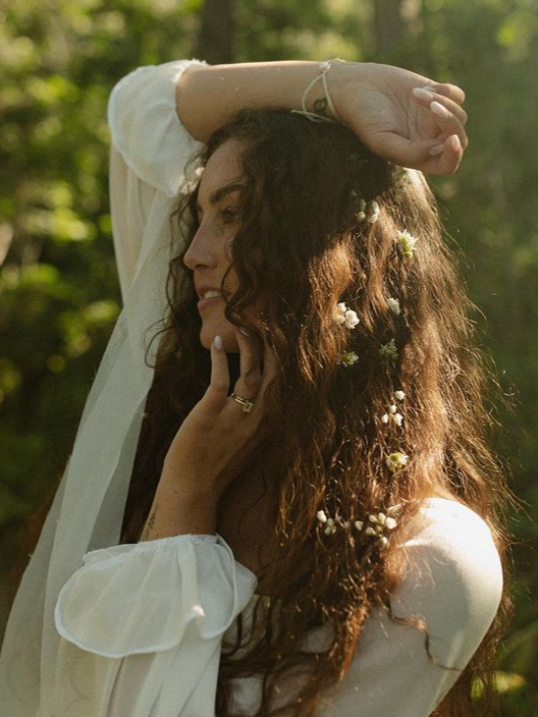 Woman with curly hair adorned with flowers, wearing a white blouse, looking away with hand raised. Outdoors.