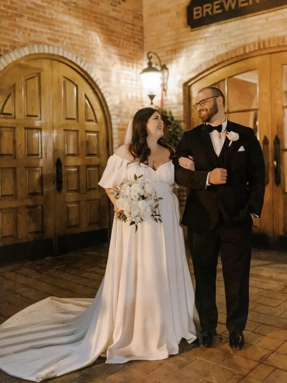 Bride and groom pose outside brewery doors. The bride wears a white gown, and the groom a black tuxedo.