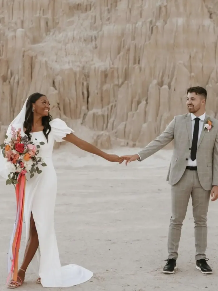 Wedding couple holding hands; woman in white dress, man in gray suit, desert backdrop.