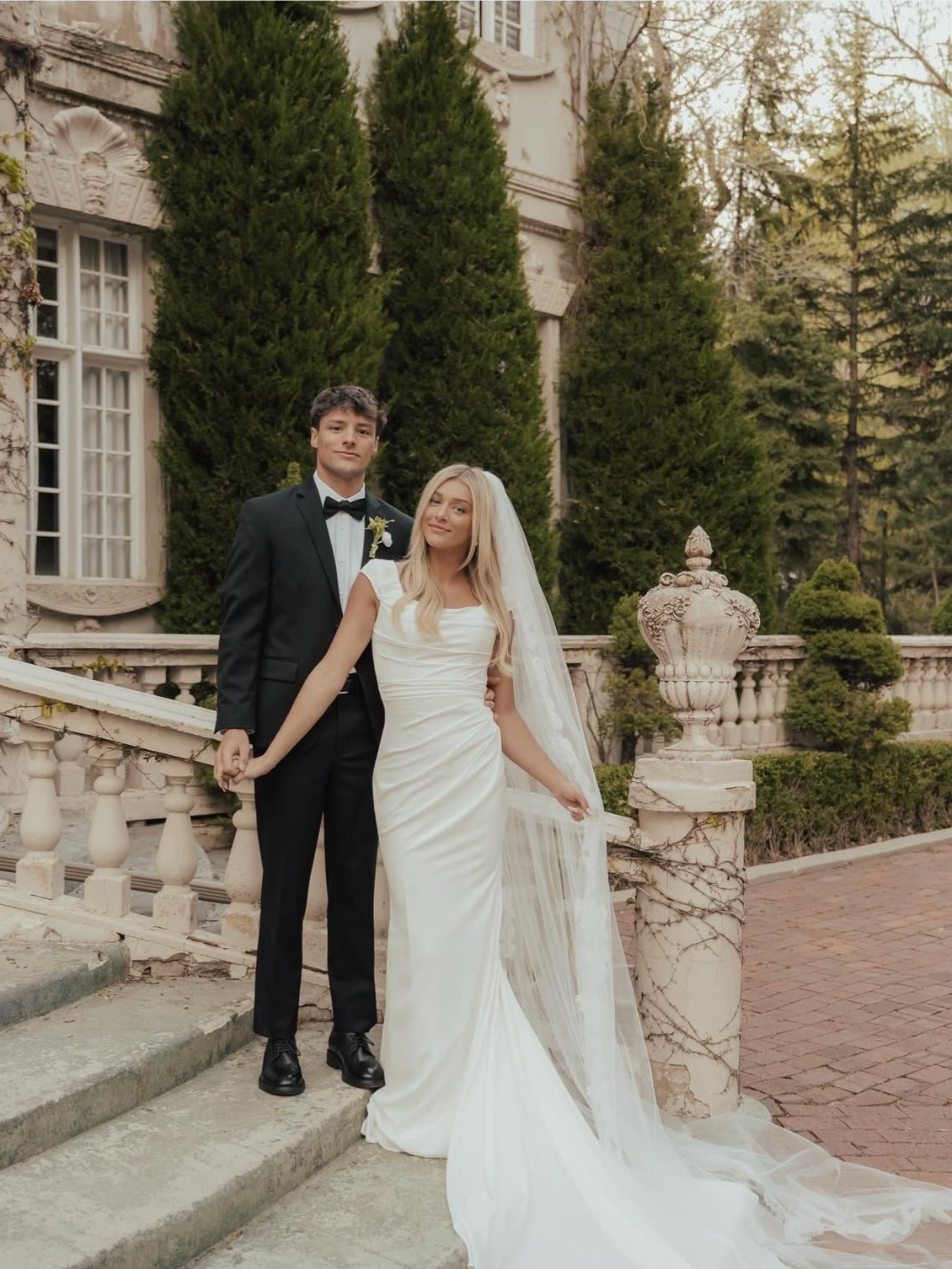 Couple on wedding day; woman in white dress, man in suit on steps in front of a building.