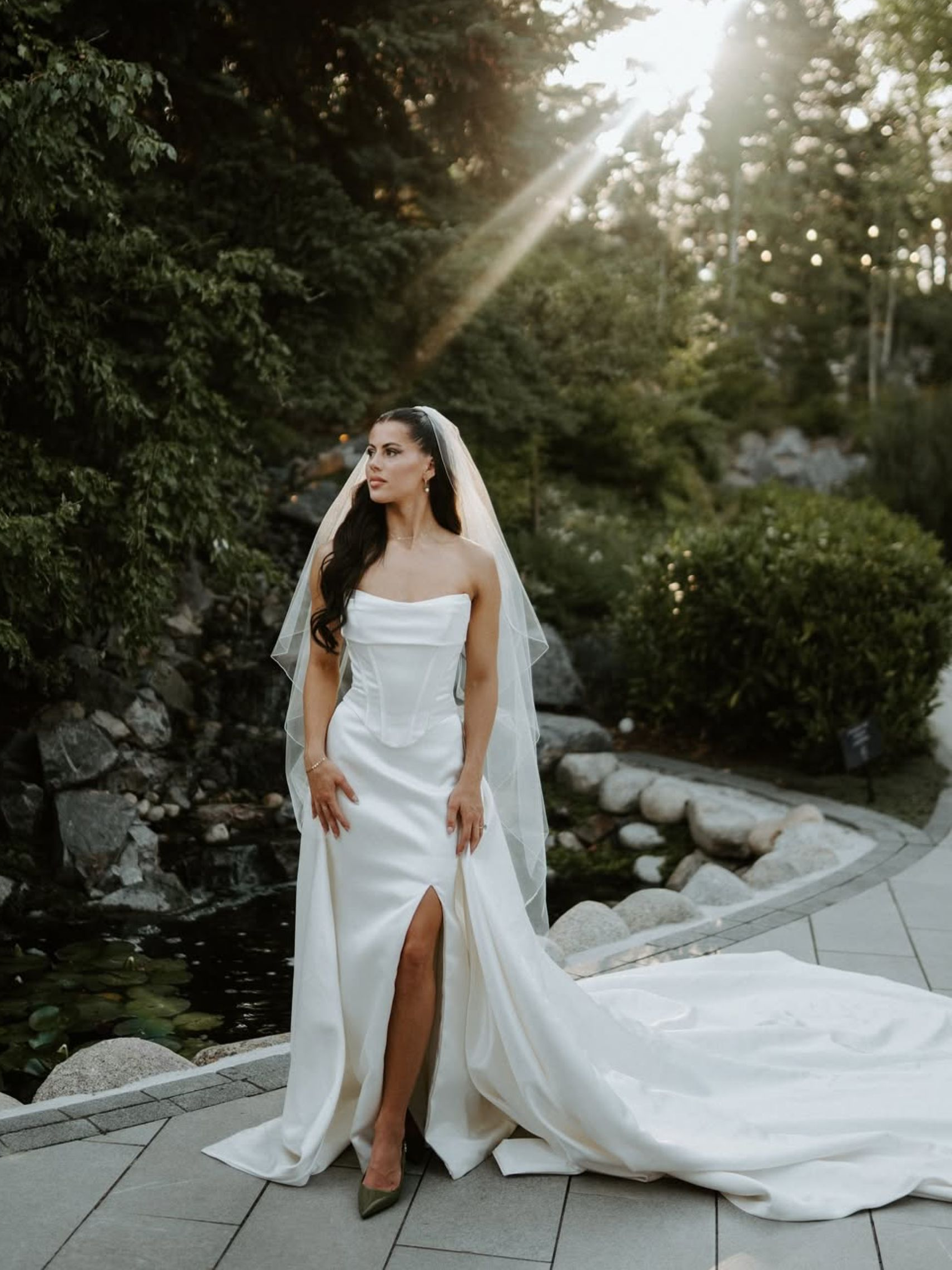 Bride in a white strapless gown with veil, standing outdoors. Sunlight streams down.