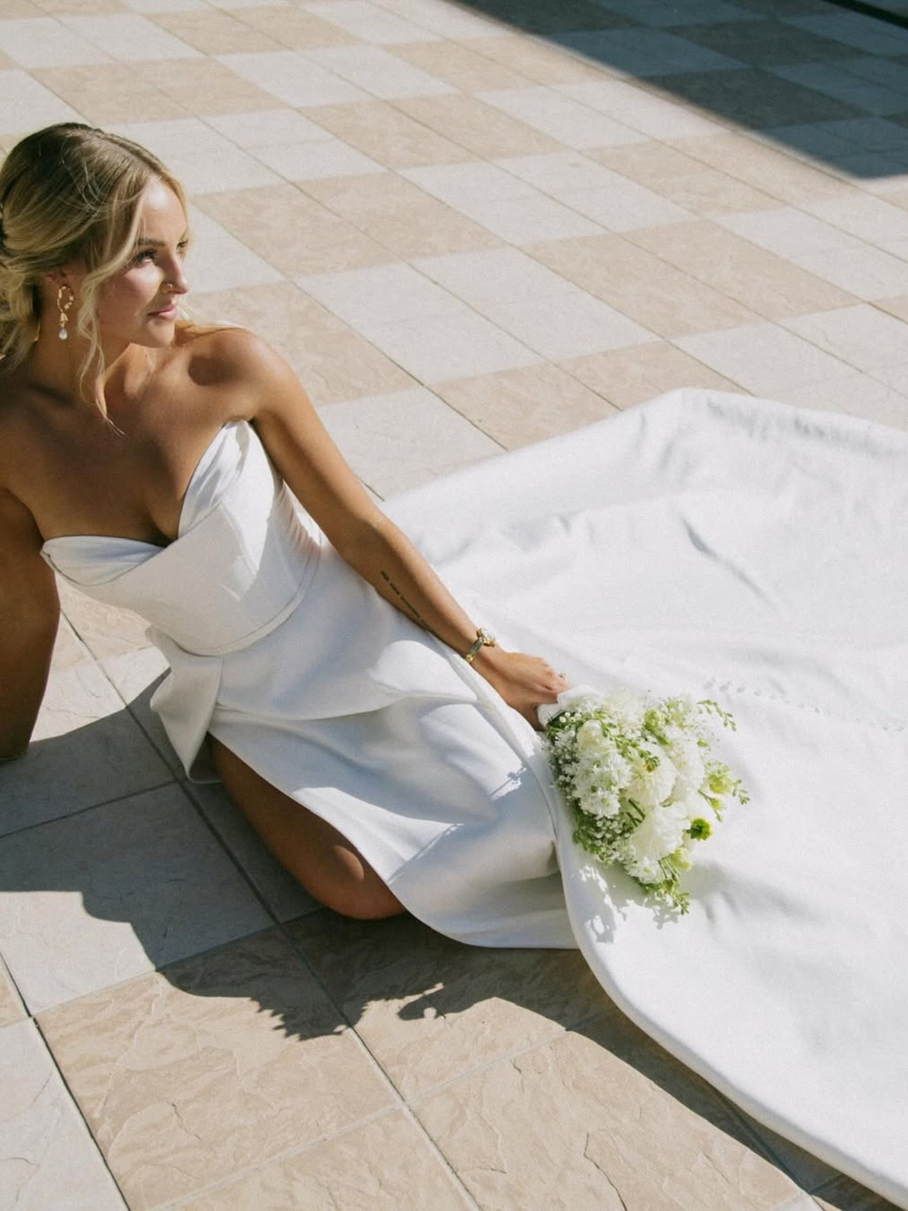 Bride in white strapless gown holding bouquet, reclining on tiled patio, looking away.