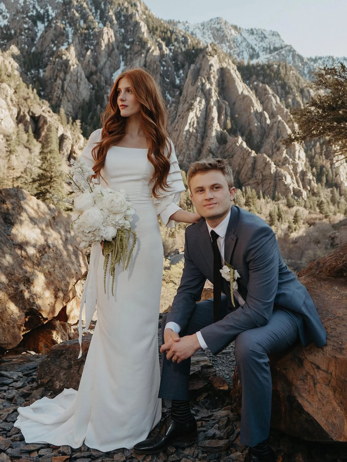 Couple in wedding attire posing outdoors with mountain backdrop. Woman holds bouquet, man sits on rock.