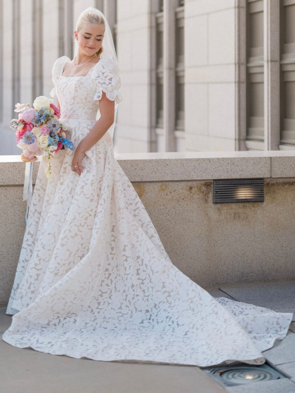 Bride in a white lace wedding dress, veil, and floral bouquet, standing outdoors.