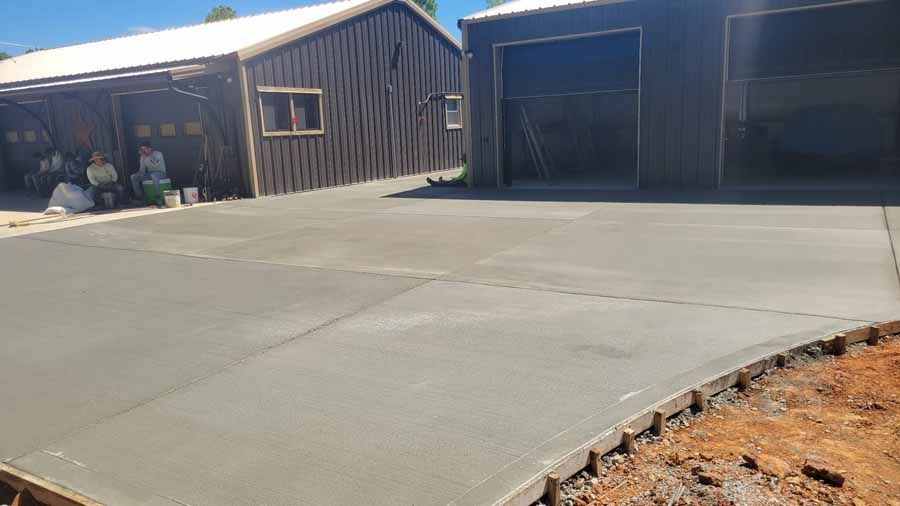 Fresh concrete driveway beside a barn-style garage and open bays, with dirt at the edge